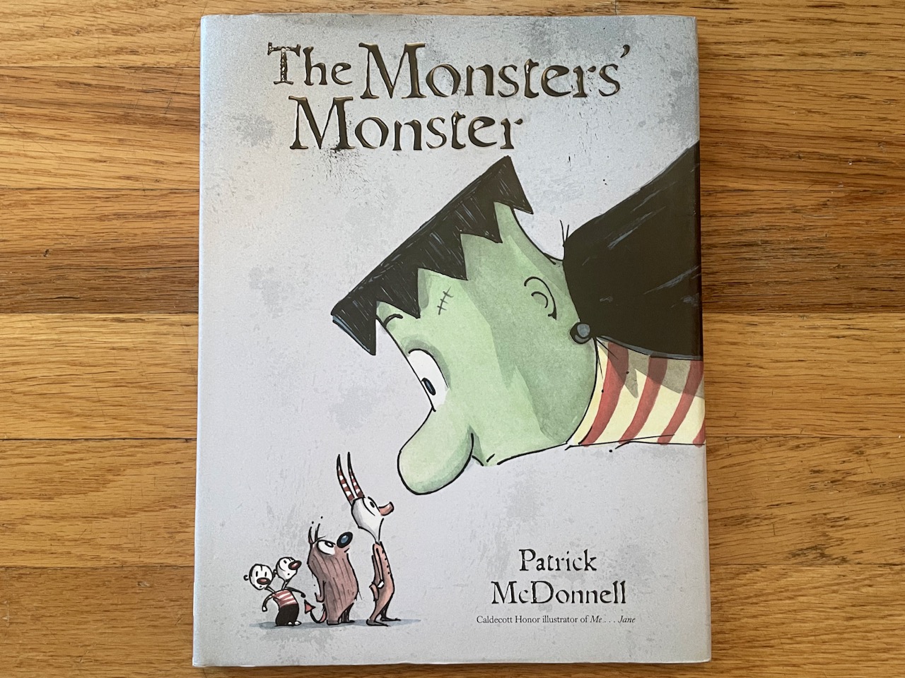 A picture book called The Monsters' Monster by Patrick McDonell. Has a large Frankenstein type monster leaning over and looking at four little monsters.